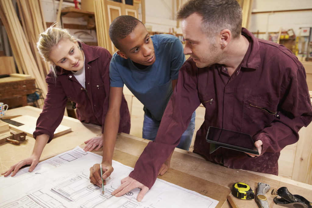 Carpenter With Apprentices Looking At Plans In Workshop | Whitehouse Communications
