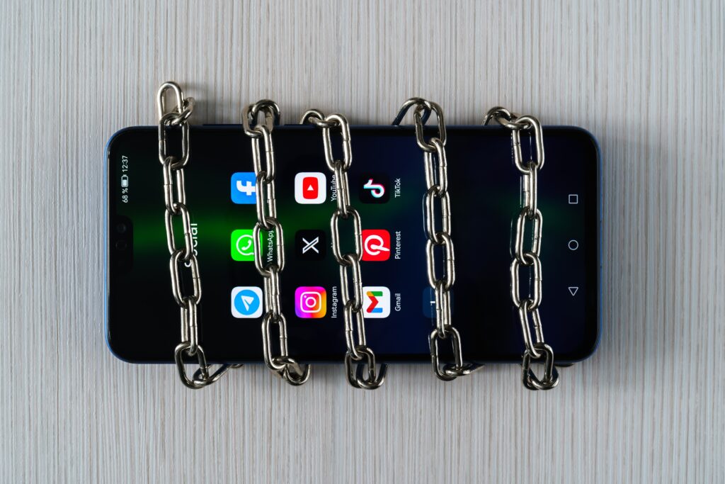 UzbekistanTashkent-January272024ChainOnAMobile | Whitehouse Communications Smartphone wrapped in sturdy metal chains on a light wooden table, conveying a locked or restricted device scenario.