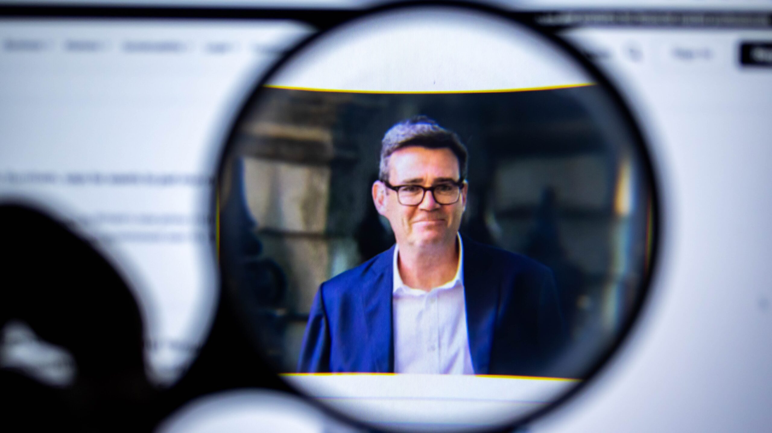 Man in a blue suit and glasses, shown on a computer screen through a magnifying glass.