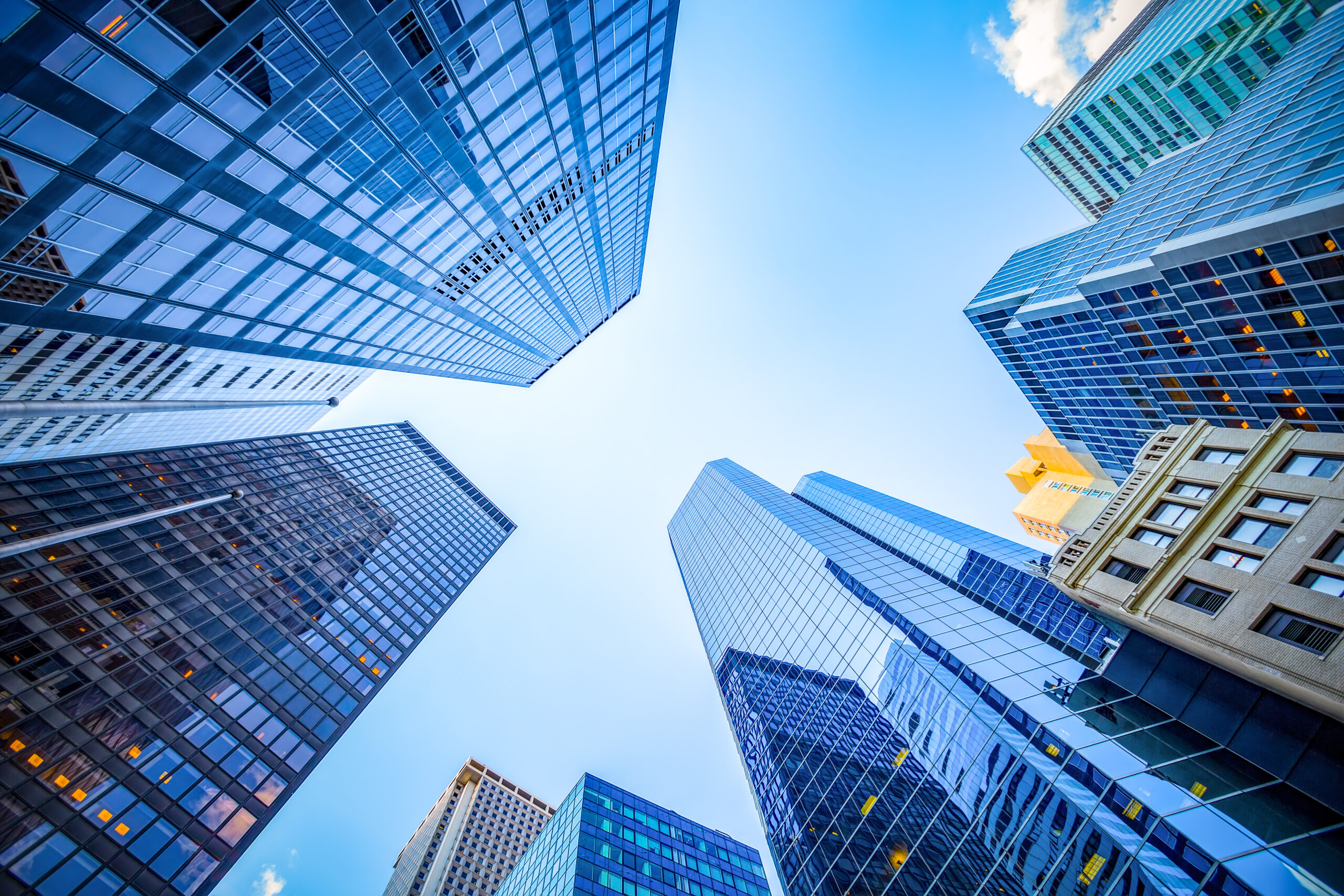 Tall glass skyscrapers converge toward a blue sky, photographed from street level looking up.